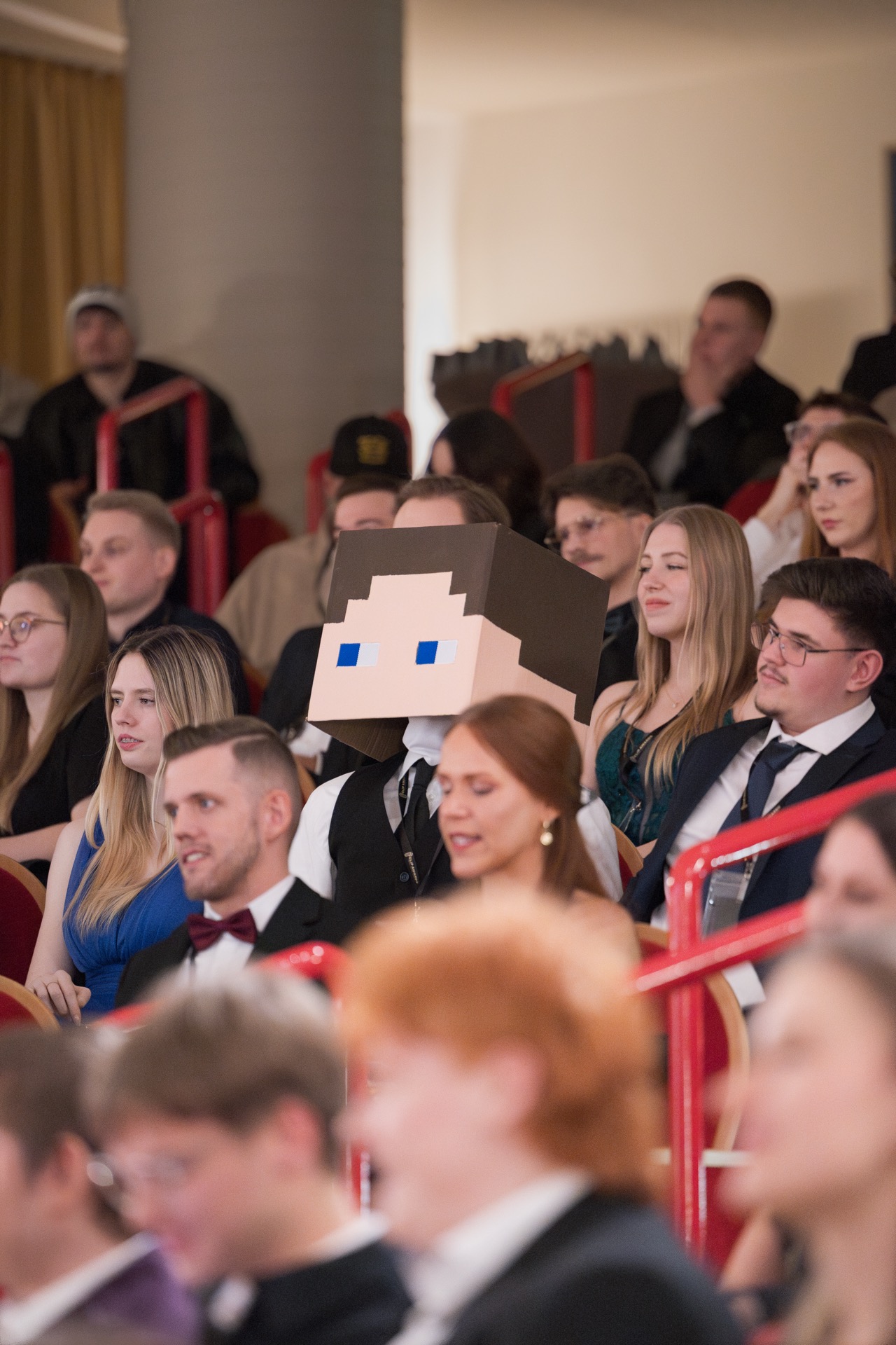 Ein Gast mit eckigem Pixelkopf sitzt mitten im Publikum
